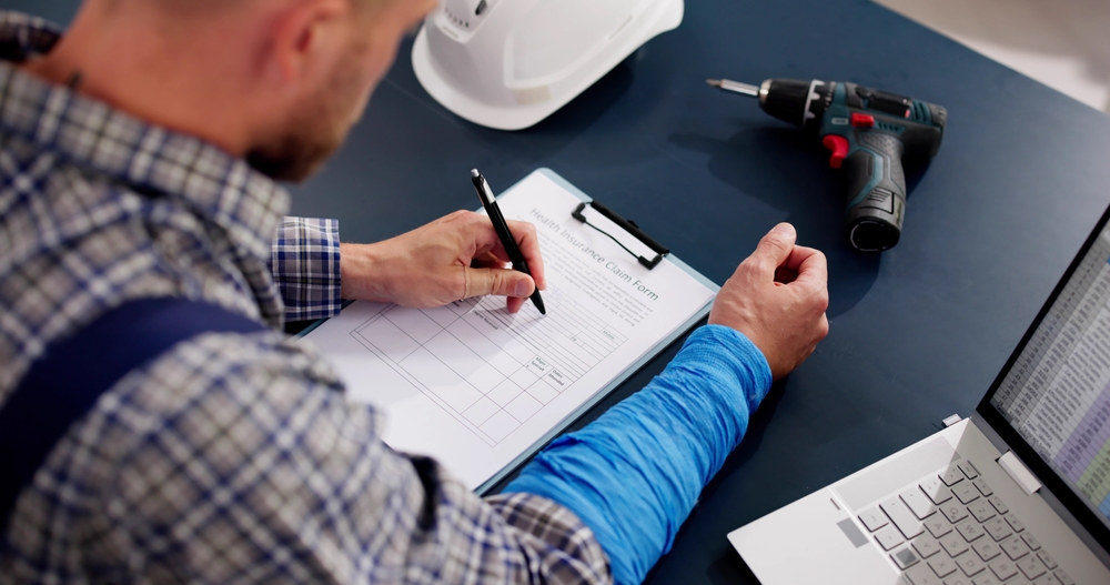 Injured worker completing a compensation form at a desk beside a laptop and tools.